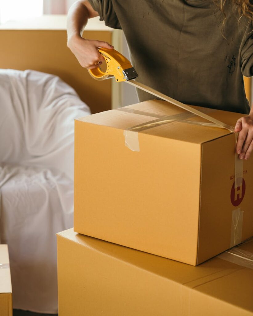 Crop female wearing casual clothes standing while packing carton boxes with scotch tape in modern apartment during preparing for relocation